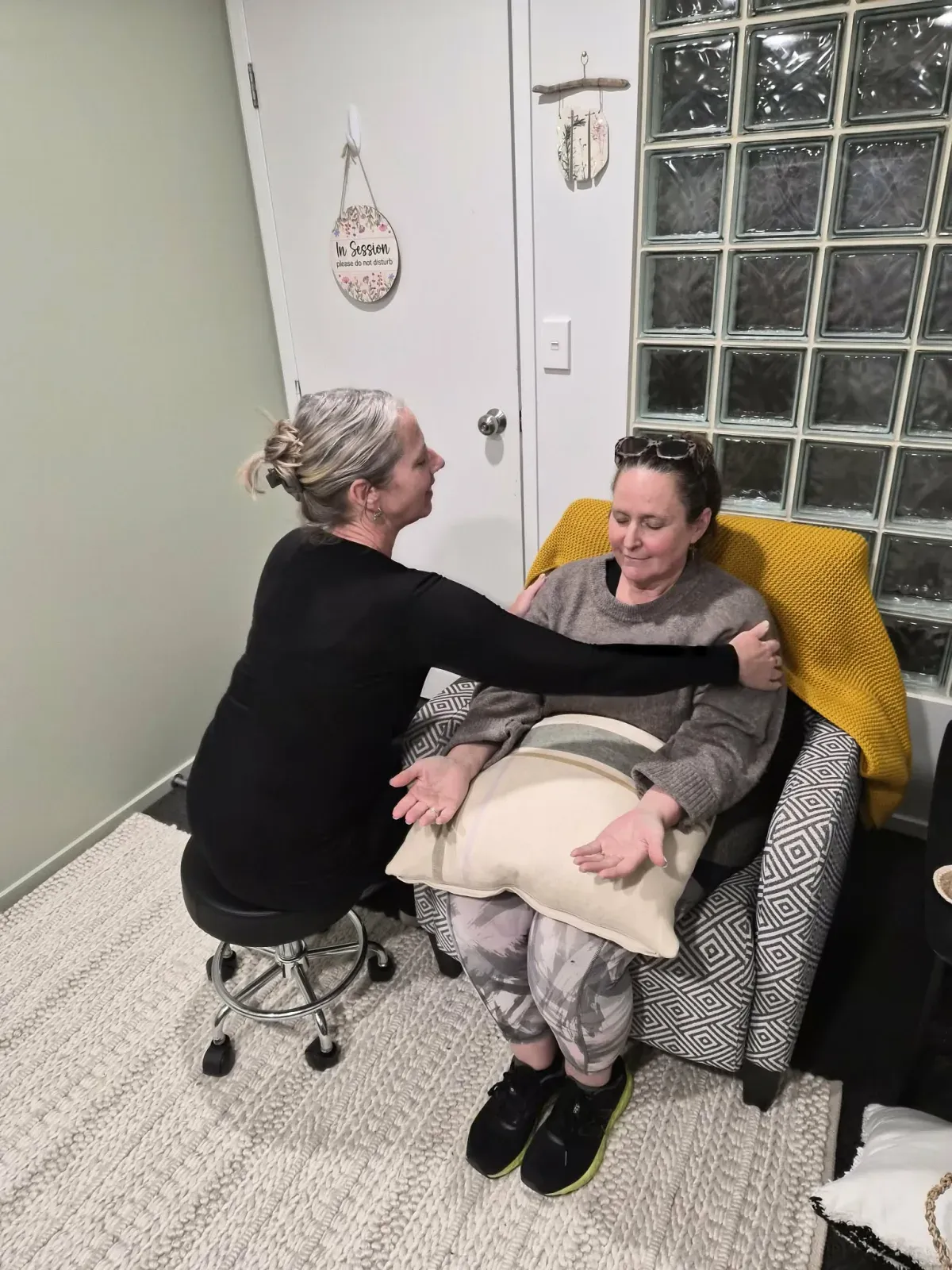Lisa seated on a low stool in the session room, gently stroking down the upper arms of a client who is settled into the armchair with her eyes closed.