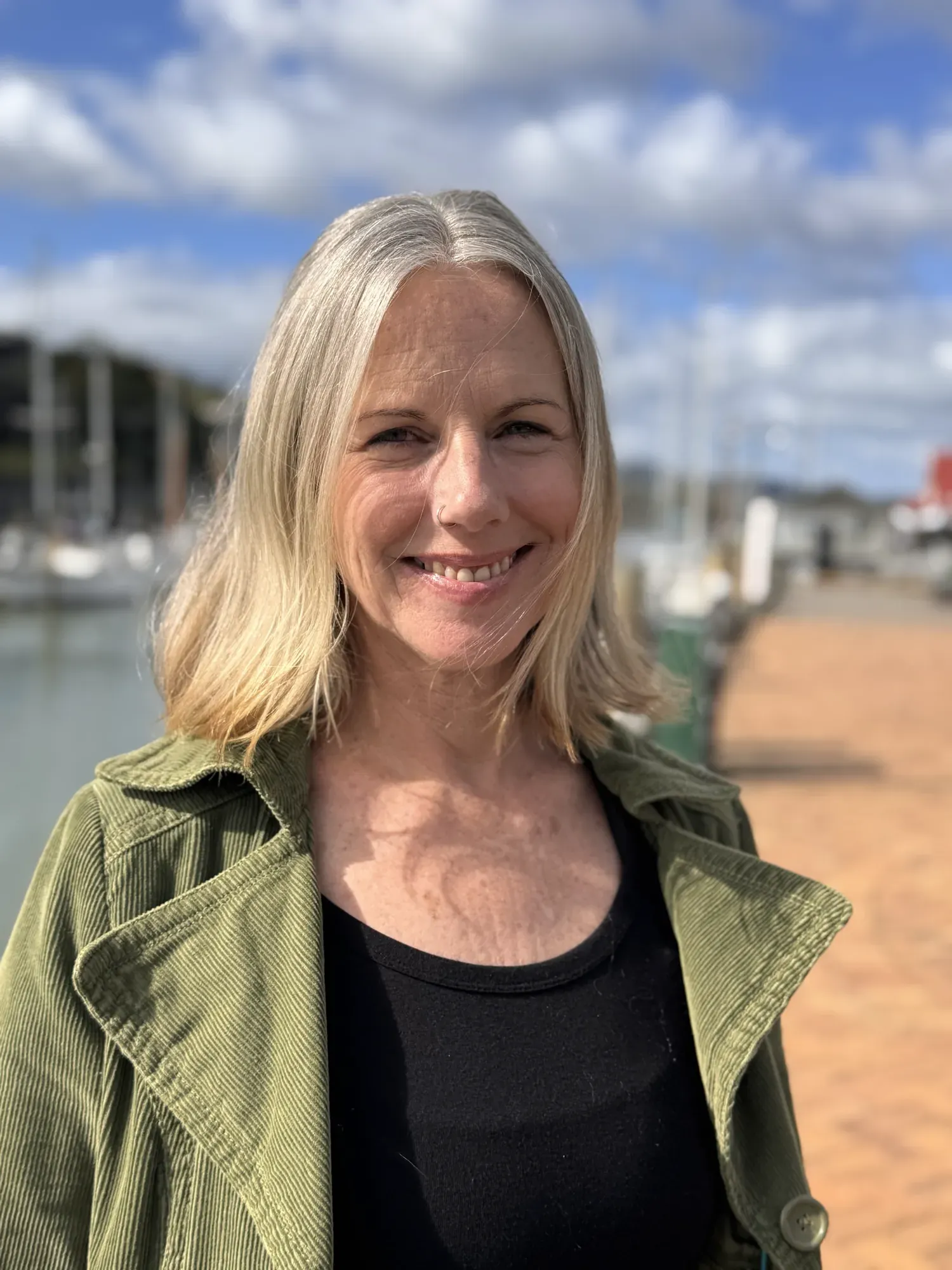 Lisa Legg, wearing a green corduroy jacket over a black top, standing on a jetty with moored boats behind her.